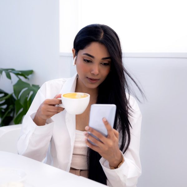 Young Indian ethnicity woman with long dark hair using smart phone and drinking coffee, lifestyle